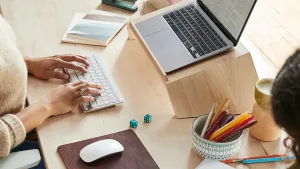 Zwei Personen arbeiten an einem Holzschreibtisch mit Laptop, Tastatur, Maus, Notizblöcken, bunten Bleistiften und Würfeln.